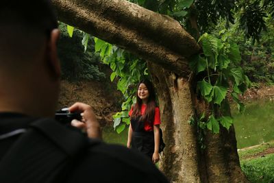 Man photographing woman standing by tree trunk