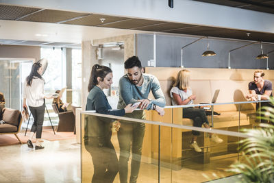 Male and female professionals talking against railing while colleagues working in background