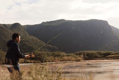 Side view of man fishing in lake against mountains and sky