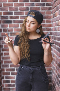 Young woman holding mobile phone while standing against brick wall