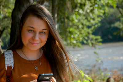 Portrait of smiling young woman holding mobile phone against trees