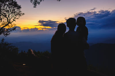 Rear view of silhouette family sitting on mountain against sky during sunset
