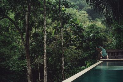 Side view of woman standing by swimming pool and wall against trees