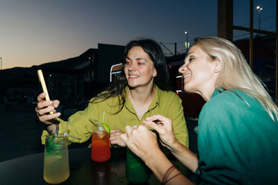 Two happy women sitting at a table in a bar drinking cocktails and chatting.