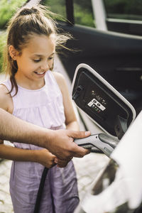 Hand of father teaching daughter to plug cable in electric car