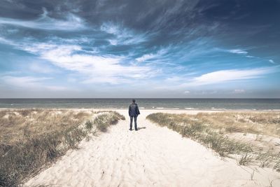Rear view of man standing on beach against cloudy sky