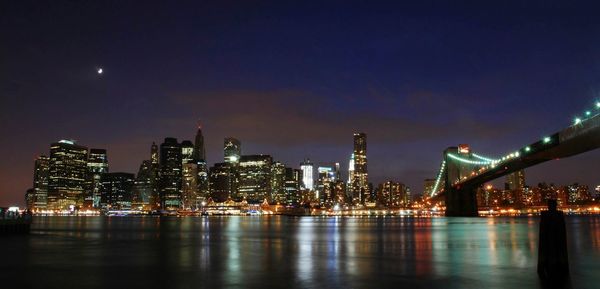 Low angle view of illuminated brooklyn bridge over river against buildings and sky in city at night