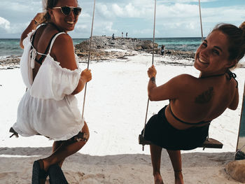 Cheerful of friends sitting on swing at beach against sky