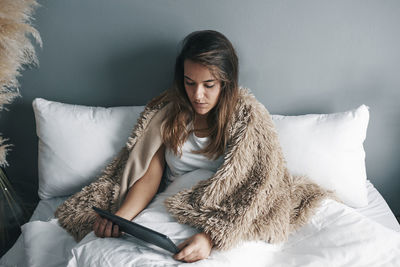 Sick young woman using digital tablet on bed against wall at home