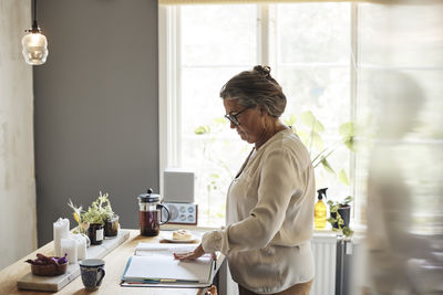 Side view of woman examining financial bills while standing in kitchen at home