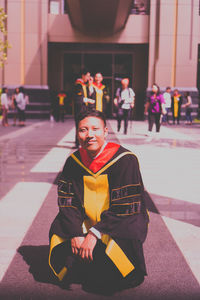 Portrait of smiling man in graduation gown crouching against building