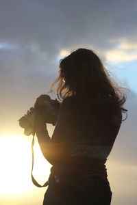 Rear view of woman holding digital video camera while standing against sky during sunset