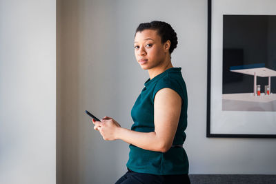 Side view of businesswoman text messaging on smart phone while standing by wall at office