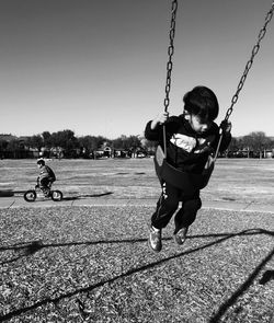 Boy siting on swing with friend riding bicycle in background against clear sky
