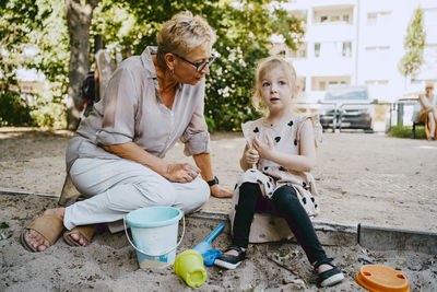 Grandmother talking to granddaughter sitting in sandbox while playing at park