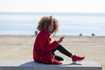 Side view of woman using smart phone while sitting on retaining wall at beach against sky