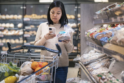 Young woman using smart phone while holding bread package and shopping in supermarket