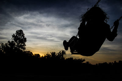 Low angle view of silhouette girl swinging on swing at park against sky during sunset