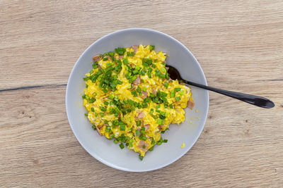 Scrambled eggs with chives and pieces of ham, lying on a plate with a fork on a wooden table. 