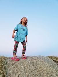 Full length of girl standing on hay bale against clear sky