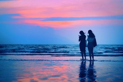 Silhouette female friends standing on shore at beach against sky during sunset