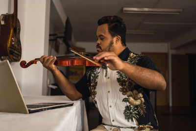 Serious young ethnic guy in casual outfit playing violin while sitting near table with netbook in light room