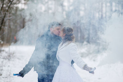 Close-up of couple with distress flare kissing against tree during winter