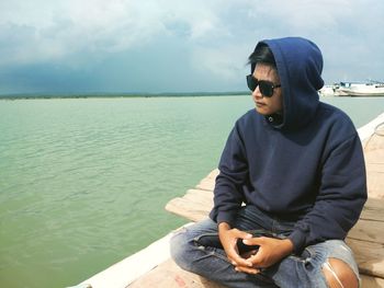 Young man sitting by sea against sky