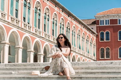 Portrait of beautiful young woman with long dark hair, sitting in square with beautiful architecture