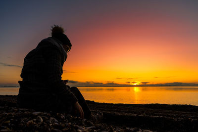 Man sitting on beach against sky during sunset
