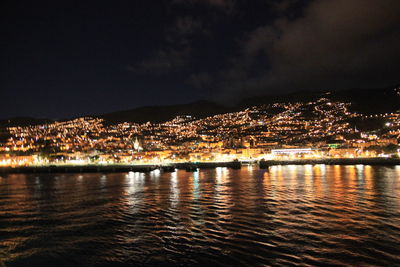 Illuminated city by sea against sky at night