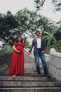 Low angle portrait of pregnant woman holding hands with boyfriend on staircase against trees