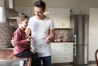 Smiling father showing smart phone to son with food while standing by kitchen counter