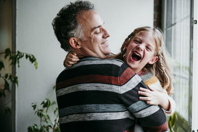 Smiling father carrying playful daughter while standing by window at home