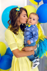 Happy mom in a yellow dress holds a young son in blue clothes , behind fly balls yellow, blue