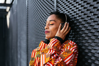 Side view of young thoughtful ethnic woman in colorful trendy shirt touching wireless headphones and listening to music with closed eyes while leaning on gray wall on city street