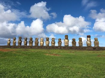 Low angle view of ancient moai statues by grassy land against cloudy sky