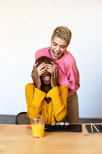 Cheerful woman covering girlfriend eyes sitting at table