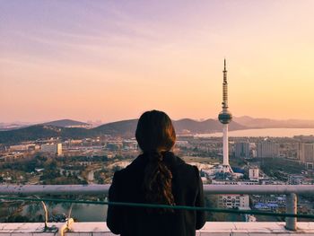 Rear view of woman standing at railing against cityscape during sunset