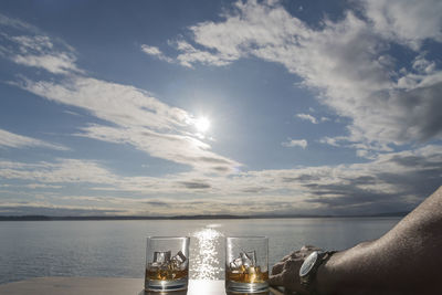 Cropped image of man hand with whiskey glasses on table by sea