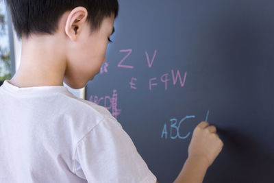 Rear view of boy writing with chalk on blackboard