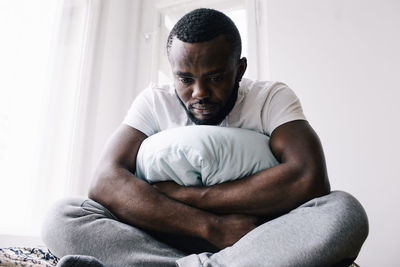 Emotionally stressed man sitting cross-legged with pillow in bedroom at home