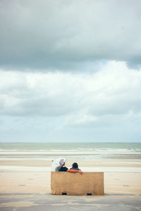Rear view of couple sitting on bench at beach against sky