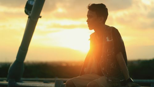 Young man looking away while sitting on railing against cloudy sky during sunset