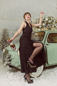 An adult woman drinks champagne at a vintage car next to a christmas tree on new year's eve