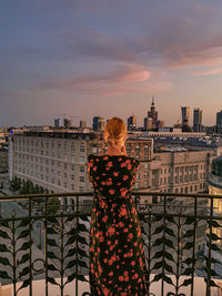 Rear view of woman standing by railing against buildings during sunset