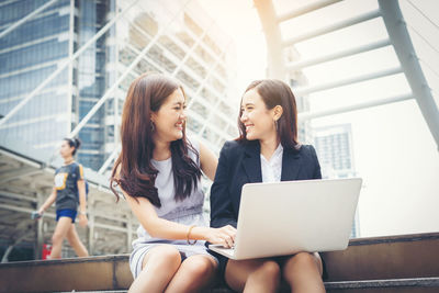 Happy young friends looking at each other while using laptop on steps in city