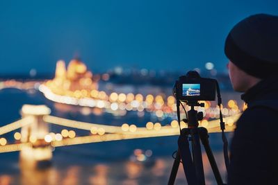 Man photographing illuminated chain bridge from camera in city at night