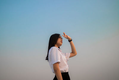 Low angle view of young woman shielding eyes standing against sky during sunset