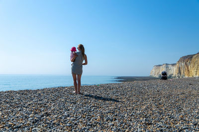 Rear view of mother carrying daughter while looking at sea against clear sky